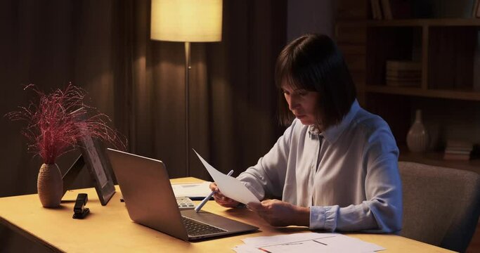 Woman continues work at home, surrounded by papers and laptop. The room is bathed in the soft glow of lamp, illuminating focused expression. With a pen in hand and eyes fixed on the screen.