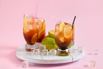 Light wooden board with glasses of tasty Long Island iced tea, ice cubes and limes on pink background