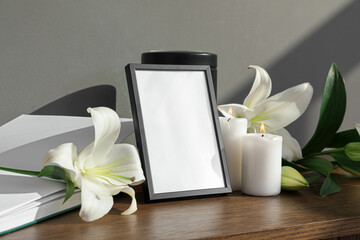 Blank funeral frame, book and white lily flowers on wooden table