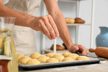 Handsome young man sprinkling fresh raw buns with sesame seeds at table in bakery