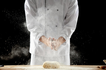 Young chef clapping and sprinkling flour over dough on table against black background