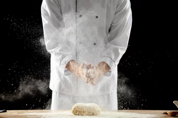 Poster Young chef clapping and sprinkling flour over dough on table against black background © Pixel-Shot