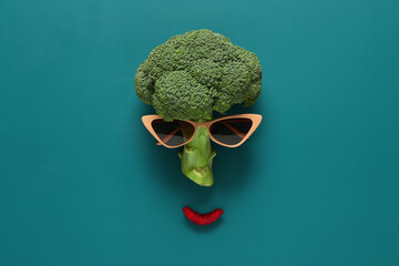 Funny face made of fresh broccoli and sunglasses on color background