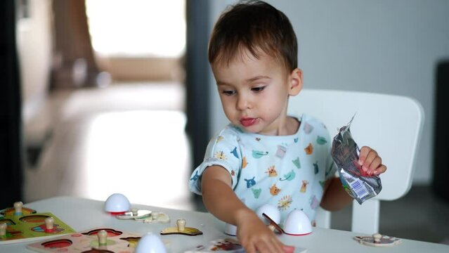 Beautiful toddler sitting at desk looking at toys. Kid drinks juice from a doypack. Blurred backdrop.