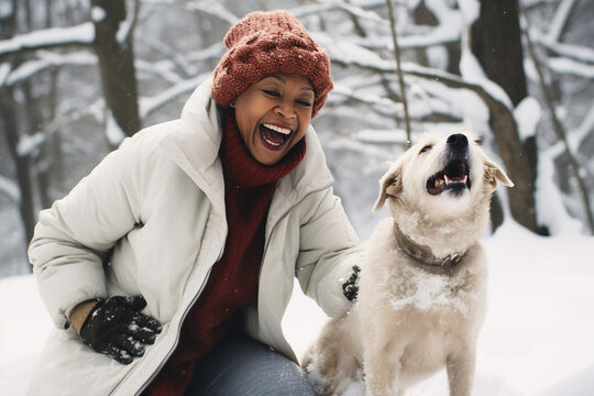 A senior african american woman is playing happily with the dog in the snow with a winter coat and winter hat in a in snow covered forest during day in winter while snowing