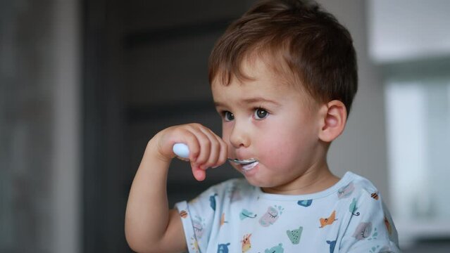 Lovely baby eating dairy from spoon himself. Calm focused child having yoghurt. Close up. Blurred backdrop.