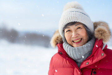 Fototapeta na wymiar A senior asian woman is playing in the snow happily with a winter coat and a winter hat in a in snow covered country landscape during day in winter while snowing