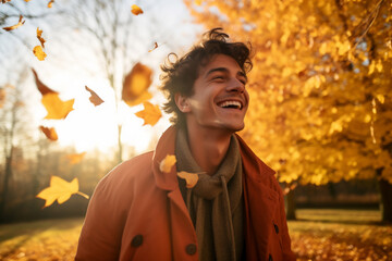 A young latin man is playing with fallen leaves happily with an autumn coat in a country landscape during sunset in autumn with a vibrant coloration