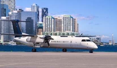 Modern white turboprop airliner with black six-bladed propellers sitting on the tarmac of Toronto City Airport separated by a stretch of blue waters of Toronto harbor from Toronto downtown with its