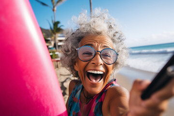 A senior latin woman is is making a selfie while smiling with a telephone on a beach while holding a surfboard a high tech social media woman