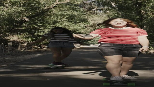 Slow motion tracking shot of girls riding skateboards in park posing for cell phone selfies  - vertical video / Saratoga Springs, Utah, United States