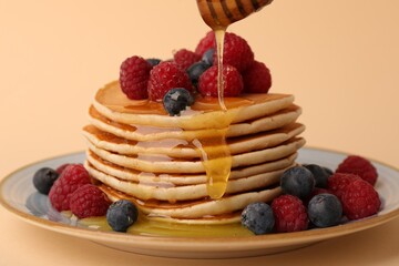 Pouring honey onto stack of tasty pancakes with raspberries and blueberries on pale orange background, closeup