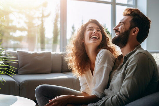 A young couple smiling brightly while sitting on a sofa by a window in a bright living room with warm light coming in - Generative AI