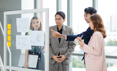 Asian professional successful young female businesswoman presenter lecturer in formal business suit outfit standing pointing color sticky note on glass board discussing with male colleague
