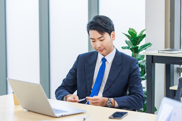 Asian professional successful male businessman employee in formal business suit sitting working typing laptop notebook in company office meeting room with graphs charts on big computer monitor screen