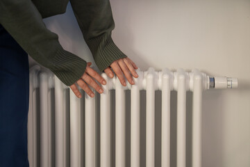 Woman warming hands near radiator at home after walking in cold winter weather, female touching barely warm battery during heating season, person near window checking heating system - obrazy, fototapety, plakaty