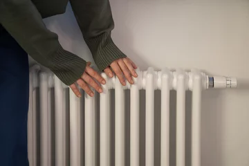 Fotobehang Woman warming hands near radiator at home after walking in cold winter weather, female touching barely warm battery during heating season, person near window checking heating system © DimaBerlin