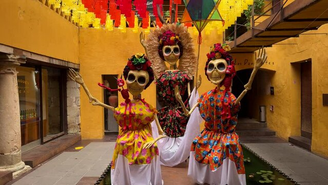 Vibrant calaveras, adorned with a riot of vivid flowers, stand gracefully in a lively Mexican courtyard. Their colorful presence embodies the spirit of Dia de los Muertos.