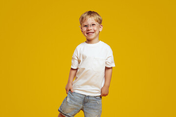 Little student boy in stylish white tshirt and eyeglasses smiling and looking at camera. Cute child laughing and standing isolated over yellow background - obrazy, fototapety, plakaty