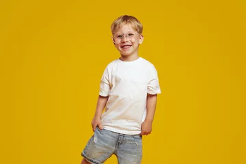 Foto op Canvas Little student boy in stylish white tshirt and eyeglasses smiling and looking at camera. Cute child laughing and standing isolated over yellow background © Davidovici