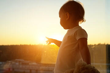 Toddler girl gazes through window holding cherished toy bear and points with a finger to sun . Child shows cuddly companion captivating sunset view outside window in hotel