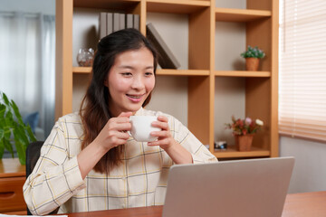 Close-up Asian woman hand holding hot coffee mug, while working in home office on laptop computer, placed in front desk, another hand is typing, working on laptop computer, happy smiling lips.