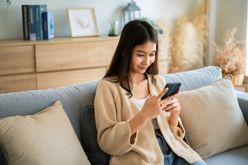 Fototapeta premium Happy young asian woman relax on comfortable couch at home texting messaging on smartphone, smiling girl use cellphone chatting, browse wireless internet on gadget, shopping online from home