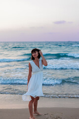 Happy woman in a white dress enjoys relaxing on the beach, walking along the sandy shore at sunset.