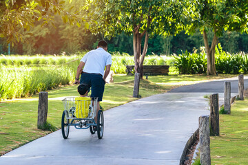 Back view of happy Asian young boy with dad ride tricycle in sunny park. Man ride tricycle with...