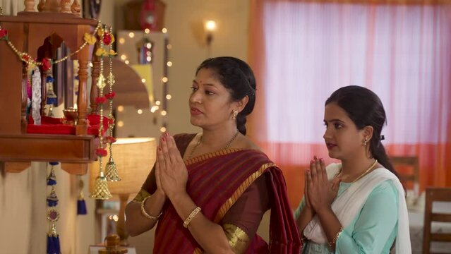 Devotional Indian Mother with daughter praying to god by folding hands in front of god mandapam at home during diwali festival celebration - concept of religious belief's, traditional culture and