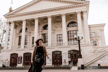 Obraz premium Woman street lifestyle. Image of stylish woman walking through European city on sunny day. Pretty woman with dark flowing hair, dressed in a beige raincoat and black, walks along the building.