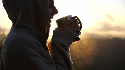 Young woman in hood meets bright sunrise and drinking hot drink Close-up of traveler enjoying hot drink from iron travel mug with steam Travel to see rising sun at sunrise envelops you in beauty. Dawn - Powered by Adobe