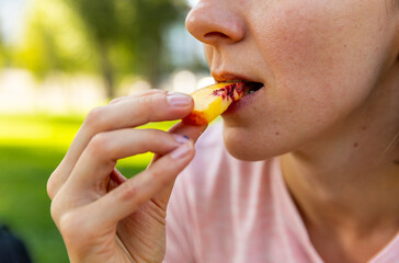 Close up of mouth eating food in public. Woman holding and bites peach or nectarine slices. Picnic concept