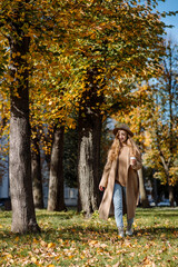 A cheerful blonde girl in a coat and hat walks in sunny windy weather in an autumn park