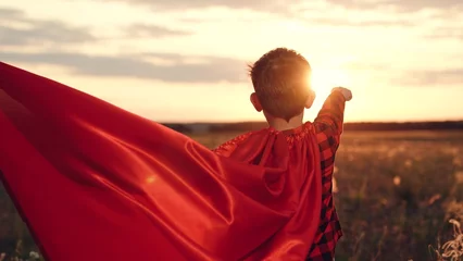 Gordijnen Playful boy stands in superhero character pose with red cape in field at sunset © SUPER FOX