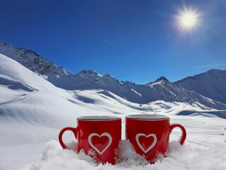 two red mug with a heart drawn on it in the snow in front of a snowy mountain landscape- winter...