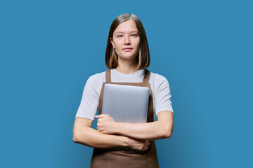 Obraz premium Portrait of young woman in apron holding laptop on blue background