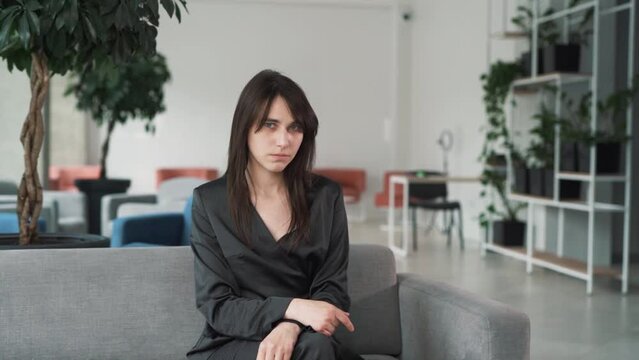 young, beautiful woman in a suit scolds while looking at the camera with a sorrowful expression on her face and gesticulates, sitting on the couch in the business center lobby