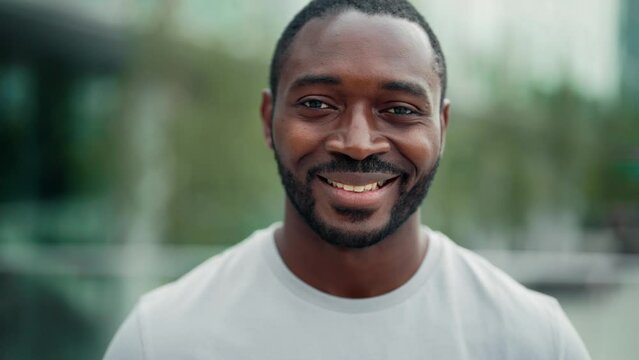 Portrait smiling positive african american bearded man on ctiy street looking at camera. Friendly charming guy on blur downtown background. Sincere human emotions, body language, good mood concept.