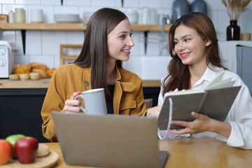 Two female students are happy while working on laptop and reading tutorial materials in home.