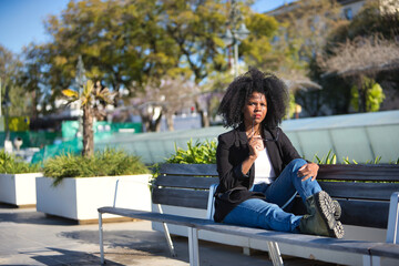 Young, beautiful, black woman with afro hair, wearing a jacket, with the temple of her glasses on her mouth, pensive, sitting on a bench. Concept thoughts, dreams, doubts, current, modern.