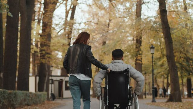 Disabled man pushing the wheels of a wheelchair with his own hands and a woman helping to push behind the back of the chair on the path in the autumn park, view from behind. Concept of acceptance