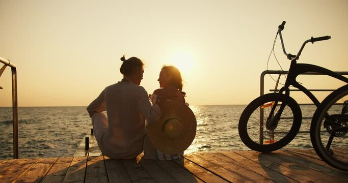 Zoom out guy and girl sitting on the pier on the beach opposite the sunrise and sea. Happy couple, a guy in light clothes and a girl with a Straw Hat, arrived on bicycles at Sunrise and are sitting