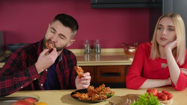 A young brunette man eats chicken wings in the kitchen at the table. A young blonde woman looks bored at it. Eating fast food. Relationships in the family.