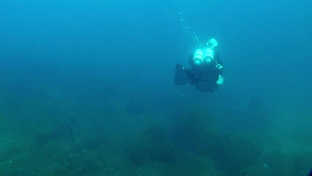 A technical diver with an underwater scooter swims over the rocky seabed, moving away from the camera.