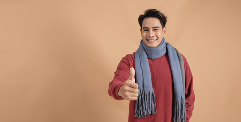 Happy and positive Asian man in red sweater and scarf giving thumbs up with ad empty space isolated...