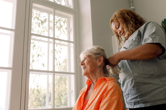 Young female caregiver braiding smiling senior woman's hair at nursing home