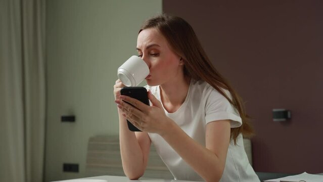 Confused woman dressed in a white T-shirt is sitting in a room, holding a smartphone in her hand, looking questioningly at a mobile phone, spreading her hand around. The girl receives unexpectedly bad