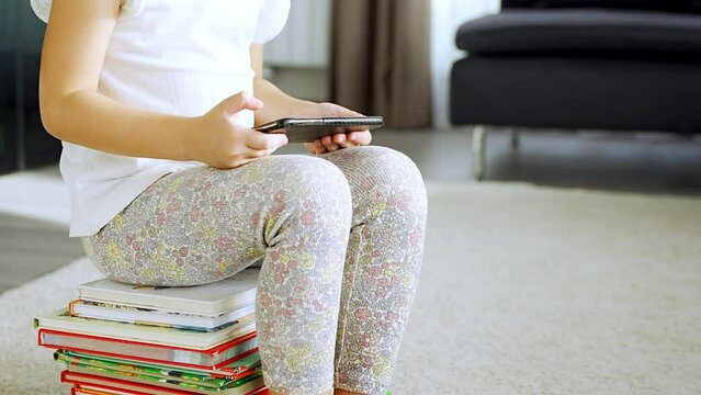 Little girl sits on stack of children's fairy-tale books and watches cartoons on her smartphone