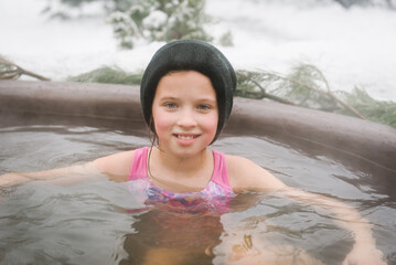 Child in hat relax in hot bath outdoors. Girl enjoying thermal spa in snowy forest. Winter holidays in mountains, hot water treatments concept. Young family in hot tub open air while snowing. Closeup.